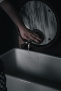 A hand is poised below a rustic, circular washbasin faucet, which is releasing a thin stream of water. The scene has a dimly lit, moody atmosphere.