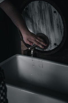 A hand is poised below a rustic, circular washbasin faucet, which is releasing a thin stream of water. The scene has a dimly lit, moody atmosphere.