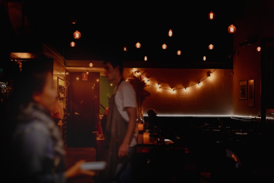 A dimly lit interior of a cozy caf&eacute; or restaurant with warm ambient lighting. Several hanging lights create a soft glow, accentuating the stylish and intimate atmosphere. A couple of people, likely patrons or staff, are visible in the foreground, slightly blurred, adding to the relaxed vibe.