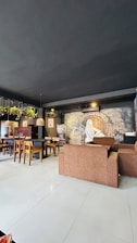 A cozy café interior features brown cushioned seats and wooden chairs around tables. The walls are adorned with artistic murals, including a figure and a brick archway. Potted plants hang from the ceiling, adding greenery to the space. Pendant lights provide warm lighting.