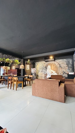 A cozy café interior features brown cushioned seats and wooden chairs around tables. The walls are adorned with artistic murals, including a figure and a brick archway. Potted plants hang from the ceiling, adding greenery to the space. Pendant lights provide warm lighting.
