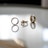 Three gold rings are positioned on a reflective surface, casting soft shadows and reflections. The first ring features a textured design, the second ring is artistically twisted, and the third is a smooth band.