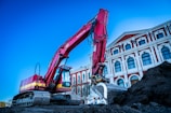A large red excavator is actively working on a construction site beside an ornate, multi-story building with classical architecture. The building has a series of arched windows and decorative elements, featuring a combination of white and red colors. The sky is clear, adding a bright backdrop to the scene.