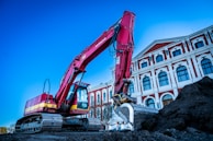 A large red excavator is actively working on a construction site beside an ornate, multi-story building with classical architecture. The building has a series of arched windows and decorative elements, featuring a combination of white and red colors. The sky is clear, adding a bright backdrop to the scene.