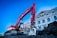 A large red excavator is actively working on a construction site beside an ornate, multi-story building with classical architecture. The building has a series of arched windows and decorative elements, featuring a combination of white and red colors. The sky is clear, adding a bright backdrop to the scene.