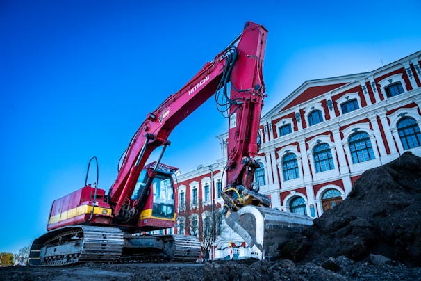 A large red excavator is actively working on a construction site beside an ornate, multi-story building with classical architecture. The building has a series of arched windows and decorative elements, featuring a combination of white and red colors. The sky is clear, adding a bright backdrop to the scene.