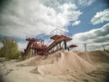 A large industrial sand quarry featuring towering red steel conveyor belts used for transporting sand. Piles of sand are scattered around, catching the sunlight. The sky is partly cloudy and contrasts with the rusted color of the machinery. Sparse vegetation is visible on the left side.
