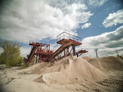 A large industrial sand quarry featuring towering red steel conveyor belts used for transporting sand. Piles of sand are scattered around, catching the sunlight. The sky is partly cloudy and contrasts with the rusted color of the machinery. Sparse vegetation is visible on the left side.