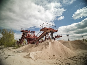 A large industrial sand quarry featuring towering red steel conveyor belts used for transporting sand. Piles of sand are scattered around, catching the sunlight. The sky is partly cloudy and contrasts with the rusted color of the machinery. Sparse vegetation is visible on the left side.