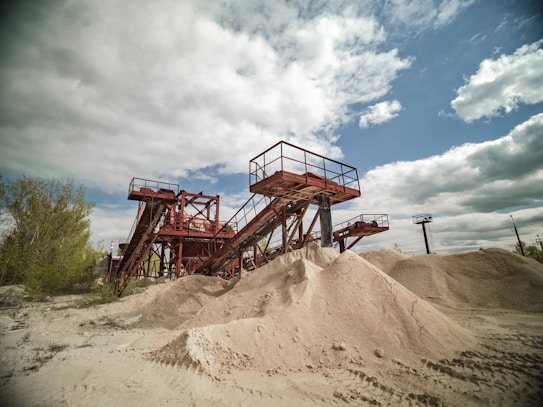 A large industrial sand quarry featuring towering red steel conveyor belts used for transporting sand. Piles of sand are scattered around, catching the sunlight. The sky is partly cloudy and contrasts with the rusted color of the machinery. Sparse vegetation is visible on the left side.
