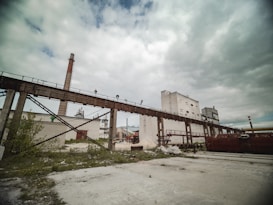 An industrial scene featuring large pipes and a long conveyor structure leading to a tall chimney. The sky is cloudy, and the ground is mostly barren with some patches of greenery. Several white buildings with minimal windows are in the background.