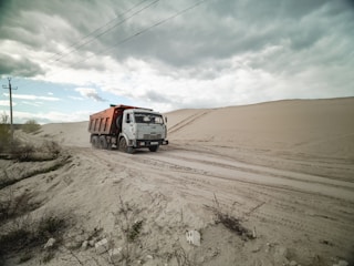 A dump truck travels along a sandy road, surrounded by large dunes under a cloudy sky. Sparse vegetation is visible in the foreground.
