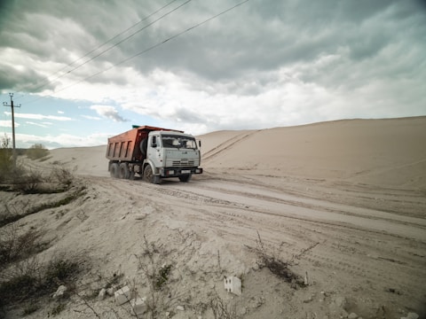 A dump truck travels along a sandy road, surrounded by large dunes under a cloudy sky. Sparse vegetation is visible in the foreground.