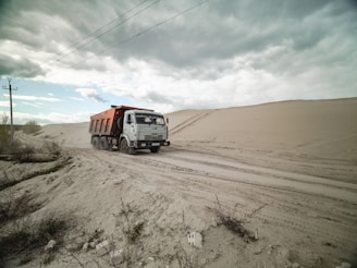 A dump truck travels along a sandy road, surrounded by large dunes under a cloudy sky. Sparse vegetation is visible in the foreground.