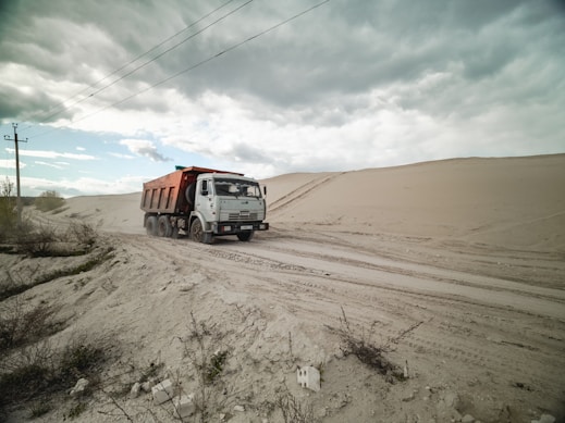 A dump truck travels along a sandy road, surrounded by large dunes under a cloudy sky. Sparse vegetation is visible in the foreground.