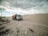 A dump truck travels along a sandy road, surrounded by large dunes under a cloudy sky. Sparse vegetation is visible in the foreground.