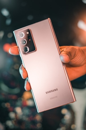 A hand is holding a sleek rose gold smartphone with a triple camera setup. The brand name Samsung is prominently visible on the back of the phone. The background is artistically blurred with circular light patterns, giving a bokeh effect.