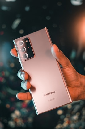 A hand is holding a Samsung smartphone with a sleek design and multiple camera lenses visible. The background is blurred with bokeh lights, enhancing a modern and tech-savvy atmosphere.