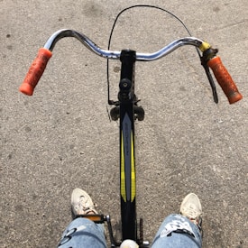 A bicycle with orange handle grips and a black frame is being ridden by someone. The person is wearing white sneakers and jeans with a design. The perspective is from above, showing the handlebars, part of the bike frame, and a concrete surface.