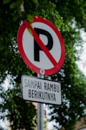 A no parking sign with a red circle and a diagonal line over the letter 'P' is mounted on a metal pole. Below it, there is a rectangular sign with Indonesian text 'SAMPAI RAMBU BERIKUTNYA'. The background is filled with green tree leaves, indicating an outdoor setting.