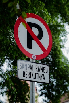 A no parking sign with a red circle and a diagonal line over the letter 'P' is mounted on a metal pole. Below it, there is a rectangular sign with Indonesian text 'SAMPAI RAMBU BERIKUTNYA'. The background is filled with green tree leaves, indicating an outdoor setting.