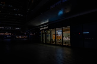A modern café store front with large glass windows revealing a brightly lit interior. Inside, there are shelves with products, a counter, and several stools for seating. The exterior is dark, casting shadows over the surrounding area but highlighting the café's sign.