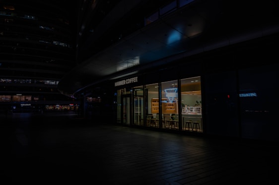 A modern café store front with large glass windows revealing a brightly lit interior. Inside, there are shelves with products, a counter, and several stools for seating. The exterior is dark, casting shadows over the surrounding area but highlighting the café's sign.