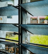 A close-up of a window with horizontal glass louvers. The glass has a textured pattern and the metal frames show signs of rust. Outside, a blurred view of a grassy area and some structures can be seen.