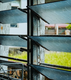 A close-up of a window with horizontal glass louvers. The glass has a textured pattern and the metal frames show signs of rust. Outside, a blurred view of a grassy area and some structures can be seen.