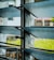 A close-up of a window with horizontal glass louvers. The glass has a textured pattern and the metal frames show signs of rust. Outside, a blurred view of a grassy area and some structures can be seen.