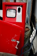 A red vintage gas pump with a handle and small sign detailing instructions is positioned in a cluttered area. The pump has a small window and metallic accents. Various objects, including what appears to be a wheel or circular part and wooden boards, surround the pump.