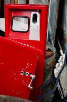 A red vintage gas pump with a handle and small sign detailing instructions is positioned in a cluttered area. The pump has a small window and metallic accents. Various objects, including what appears to be a wheel or circular part and wooden boards, surround the pump.