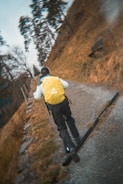 A person wearing a yellow backpack and outdoor clothing is running uphill on a gravel path. The surrounding area is grassy with some trees and the path appears to lead through a natural landscape.