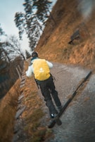 A person wearing a yellow backpack and outdoor clothing is running uphill on a gravel path. The surrounding area is grassy with some trees and the path appears to lead through a natural landscape.