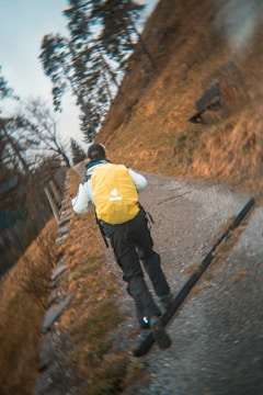 A person wearing a yellow backpack and outdoor clothing is running uphill on a gravel path. The surrounding area is grassy with some trees and the path appears to lead through a natural landscape.