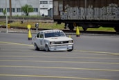 A white racing car with decals and the word 'Kadett' on the windshield is navigating around traffic cones on a marked road surface. In the background, train tracks and a large freight train with graffiti are visible, along with an industrial building.