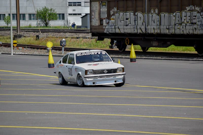 A white racing car with decals and the word 'Kadett' on the windshield is navigating around traffic cones on a marked road surface. In the background, train tracks and a large freight train with graffiti are visible, along with an industrial building.