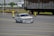 A white racing car with decals and the word 'Kadett' on the windshield is navigating around traffic cones on a marked road surface. In the background, train tracks and a large freight train with graffiti are visible, along with an industrial building.