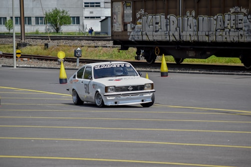 A white racing car with decals and the word 'Kadett' on the windshield is navigating around traffic cones on a marked road surface. In the background, train tracks and a large freight train with graffiti are visible, along with an industrial building.