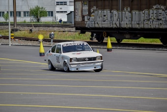 A white racing car with decals and the word 'Kadett' on the windshield is navigating around traffic cones on a marked road surface. In the background, train tracks and a large freight train with graffiti are visible, along with an industrial building.