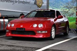 A red Honda Integra car is parked on an asphalt surface with a mountainous background. The car features sporty white wheels and distinct headlights. It's positioned near other vehicles and equipment, suggesting a car event or race preparation.
