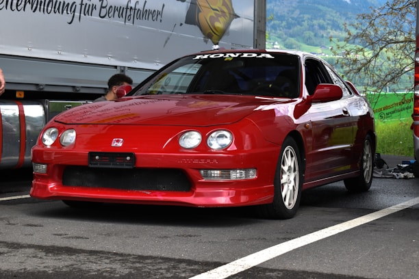 A red Honda Integra car is parked on an asphalt surface with a mountainous background. The car features sporty white wheels and distinct headlights. It's positioned near other vehicles and equipment, suggesting a car event or race preparation.