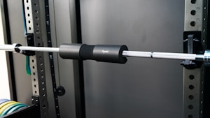 A gym setup with a metal barbell fitted with a black protective pad is secured on a squat rack. The background includes metal frames with numbered holes, suggesting adjustability for different exercises. Colored weight plates are visible on the lower left, stacked in an organized manner.