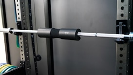 A gym setup with a metal barbell fitted with a black protective pad is secured on a squat rack. The background includes metal frames with numbered holes, suggesting adjustability for different exercises. Colored weight plates are visible on the lower left, stacked in an organized manner.