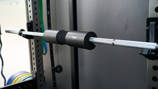 A barbell is mounted on a squat rack, equipped with a black foam pad for cushioning. The metal barbell has a textured grip, and the setup is surrounded by a gym environment with various colored weights visible in the background.