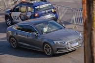 A gray Audi car is driving on a street, with a driver visible inside. In the background, there is a parked blue and white police vehicle with barriers set up around it. The scene suggests a situation involving traffic control or a checkpoint.
