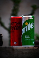 Two beverage cans are placed next to each other on a surface. The can on the left is red with black text, while the can on the right is green with white text. Both cans have condensation on their surfaces, suggesting they are cold.