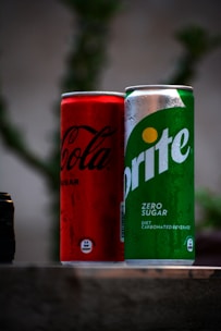 Two beverage cans are placed next to each other on a surface. The can on the left is red with black text, while the can on the right is green with white text. Both cans have condensation on their surfaces, suggesting they are cold.