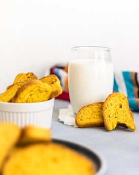 A glass of milk is placed on a light surface, accompanied by several pieces of crispy, golden-brown toast. The toast is partly in a white bowl and also scattered around. In the background, a colorful striped cloth adds a touch of color to the composition.
