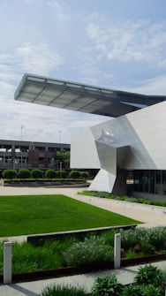 A modern architectural structure with a large overhanging design element made of metal and glass. The building has clean, angular lines and is surrounded by a well-manicured lawn and landscaped greenery. In the background, there are additional buildings and a partly cloudy sky.